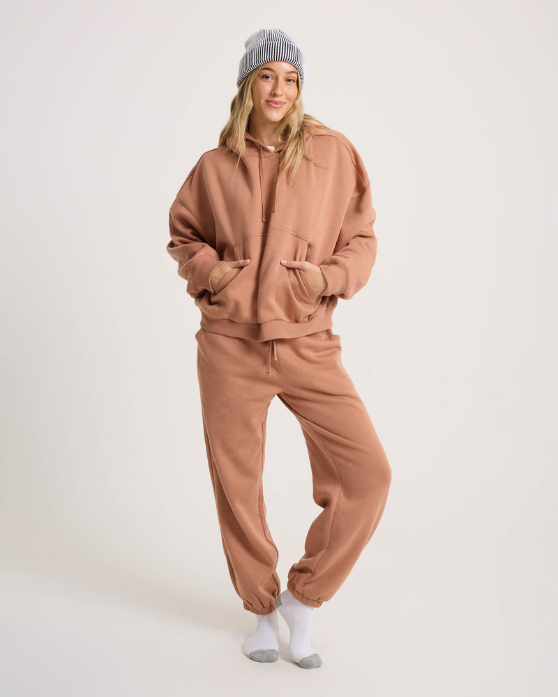 A person in an oversized tan hoodie, Volcom Women’s In A Cinch Jogger Pant, gray and white beanie, and white socks stands smiling, capturing everyday comfort against a plain white background.