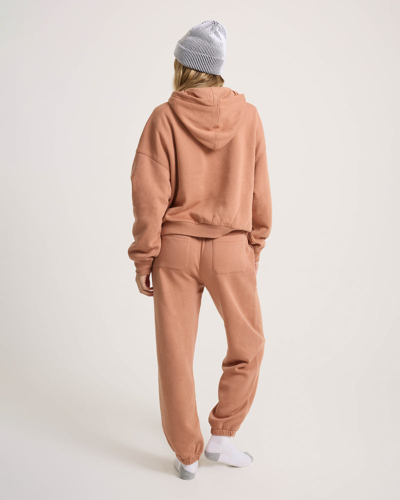 A person wearing Women’s In A Cinch Jogger Pant by Volcom, an oversized peach hoodie, white socks, and a gray knit beanie stands facing away against a plain light background, embodying everyday comfort.
