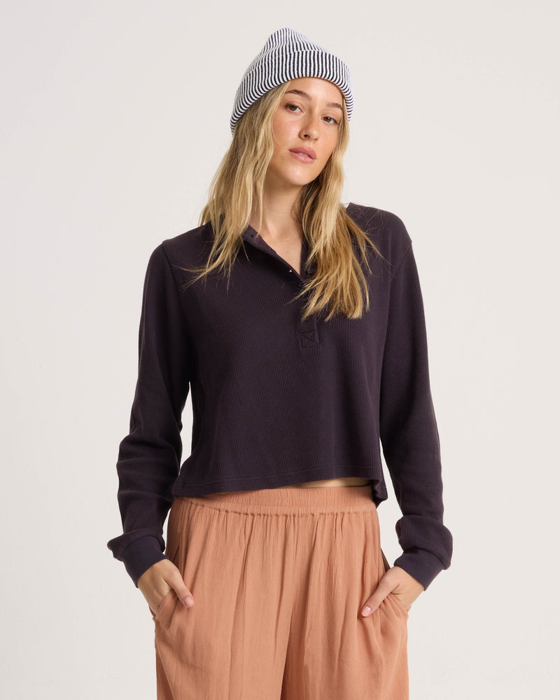 A person wears a dark long-sleeved Volcom Women’s Ski Bum Henley, peach-colored pants, and a striped knit beanie.