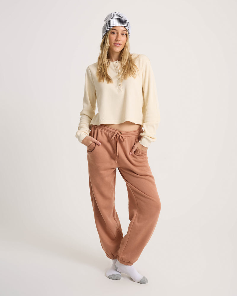 A woman wearing a Volcom Women’s Ski Bum Henley in cream, paired with brown drawstring sweatpants and a striped beanie.