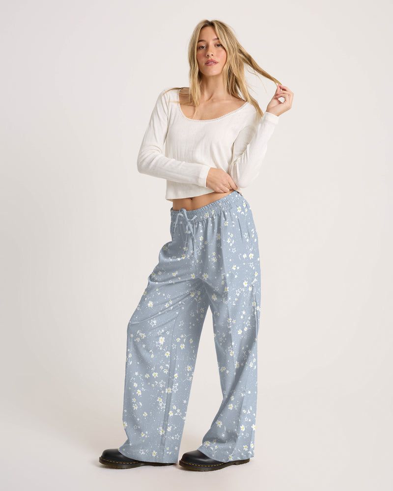 A woman wears a white long-sleeved top and Volcom Women’s Longboard Wide Leg Pant, which are loose, light blue, and feature a small white floral print.