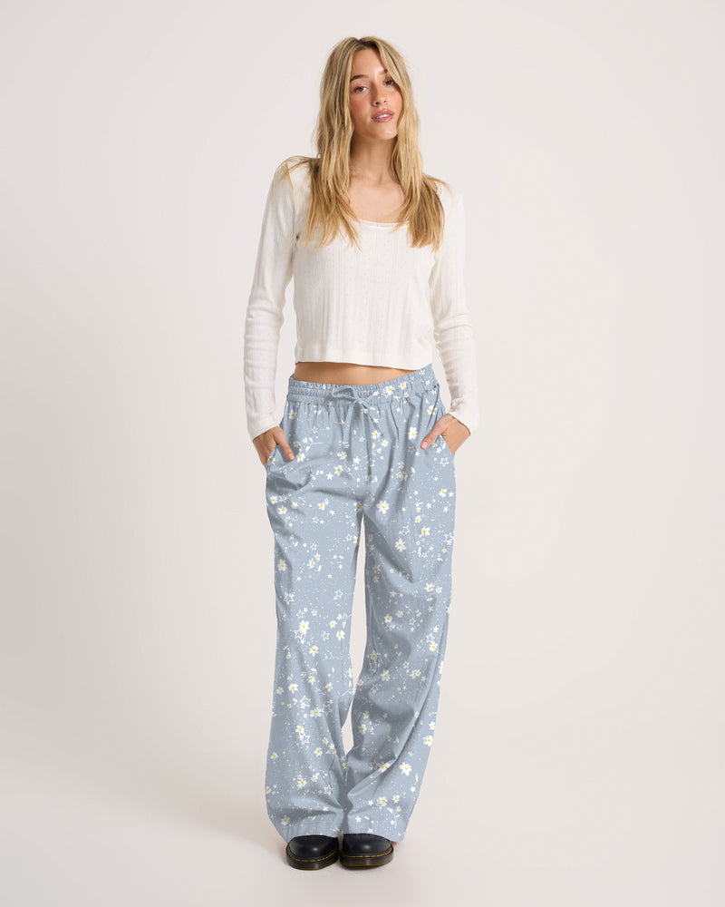 A person wears a white long-sleeved shirt and Volcom Women’s Longboard Wide Leg Pant in lightweight blue viscose with a white daisy print and a drawstring waist.
