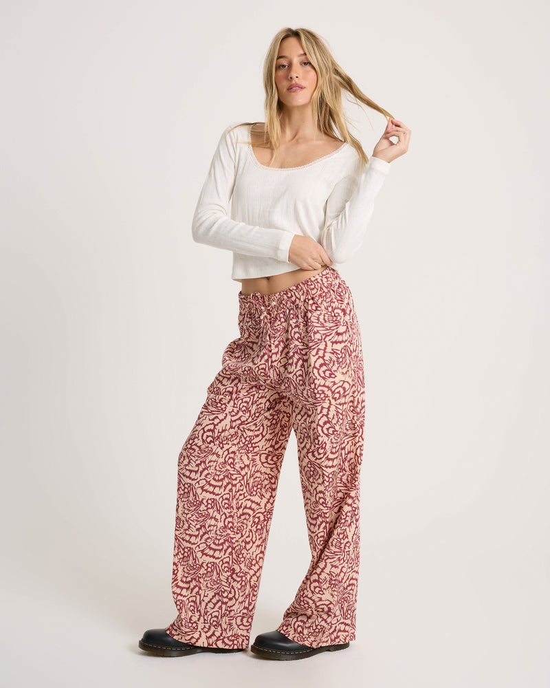 A woman wears a white long-sleeved top and Volcom Women’s Longboard Wide Leg Pant, featuring a patterned, relaxed fit and a drawstring waist.