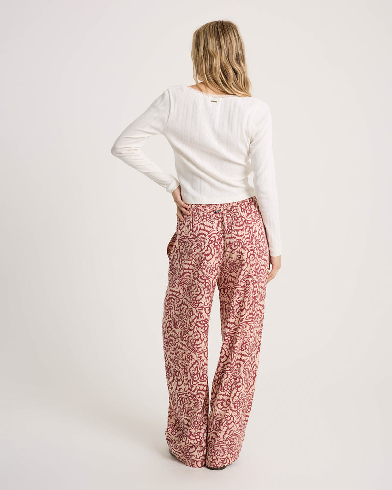 A person seen from behind wears a white long-sleeved top and Volcom Women’s Longboard Wide Leg Pant, featuring a red and cream-colored abstract print with a relaxed fit.