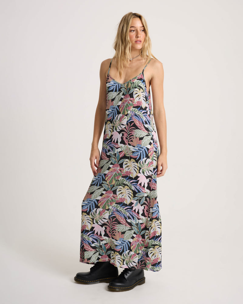 A woman wears a floor-length tropical leaf print Volcom Women’s Cosmic Slip Dress with black lace-up boots.