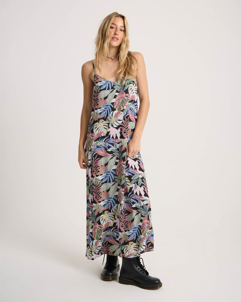 A woman wearing a Volcom Women’s Cosmic Slip Dress featuring a black spaghetti-strap maxi design with a colorful tropical leaf print, styled with black lace-up boots.