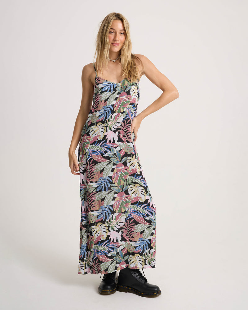 A person wears a vibrant, multicolored tropical leaf print Women’s Cosmic Slip Dress with spaghetti straps, styled with black lace-up boots.