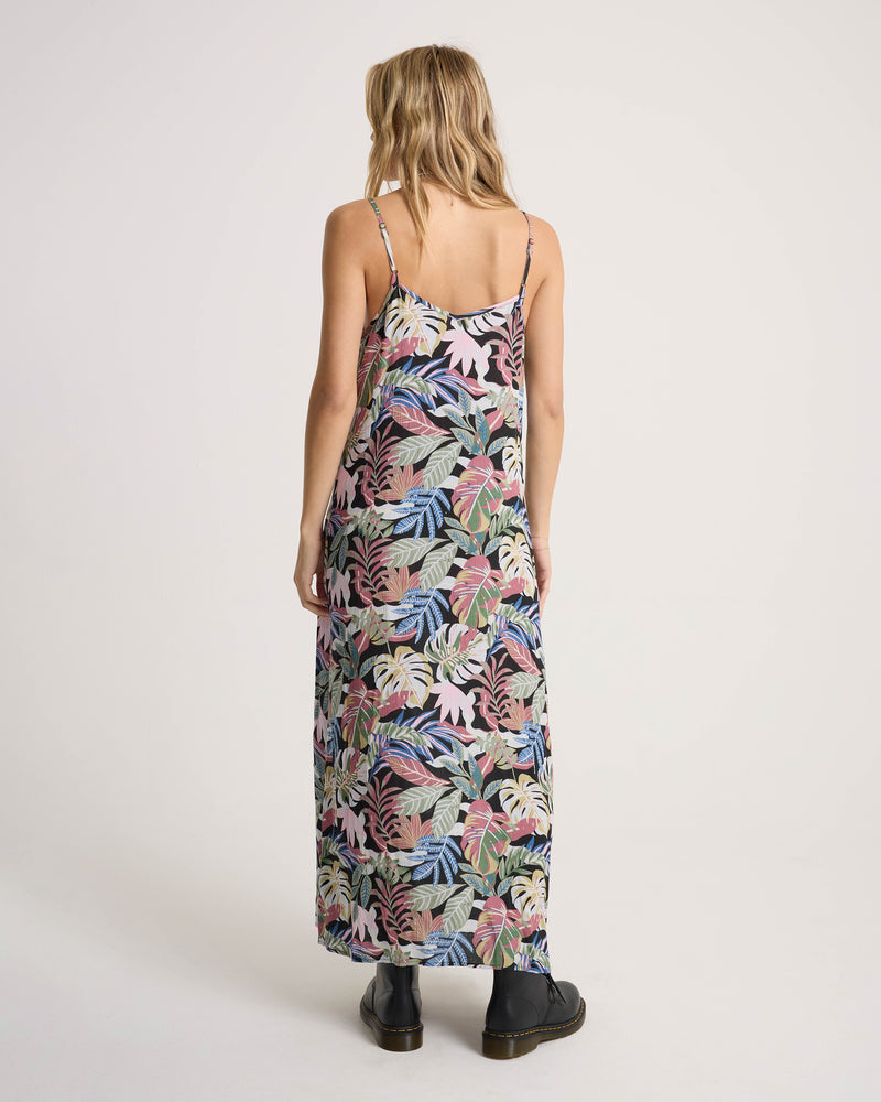 Back view of a person wearing a Volcom Women’s Cosmic Slip Dress featuring a black background with a colorful tropical leaf print, paired with black ankle boots.