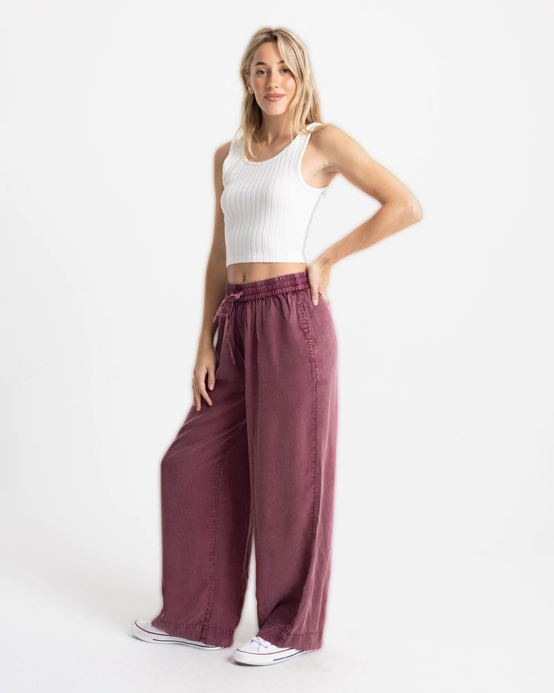 A person wearing a white ribbed tank top and Volcom Women’s Washed Up Twill Pant in maroon, featuring a loose, wide-leg fit and an elastic drawstring waist.