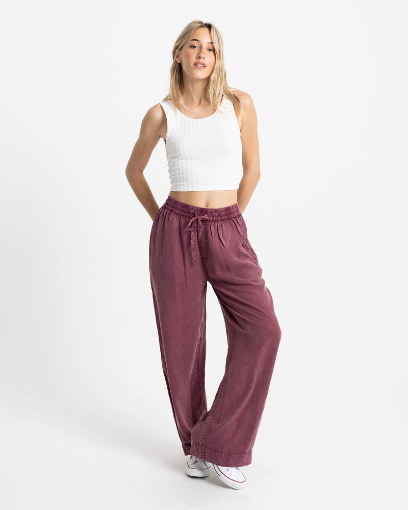 A woman wears a white ribbed tank top and Volcom Women’s Washed Up Twill Pant in maroon, featuring a wide-leg fit and a tie-front waist.