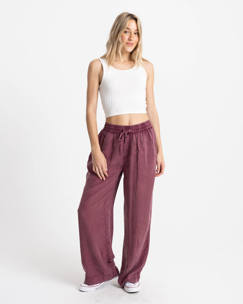 A woman wears a white ribbed tank top and Volcom Women’s Washed Up Twill Pant in burgundy, featuring a loose-fit, high-waisted design with a tie-front waist.