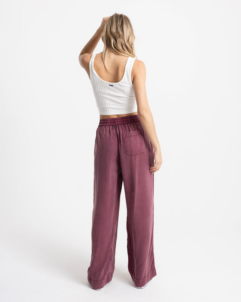 A person seen from behind wears a white tank top and Volcom Women’s Washed Up Twill Pant in burgundy, featuring a loose, wide-leg fit, an elastic waistband, and a back pocket.