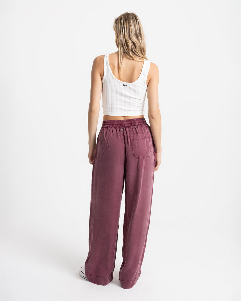 A person wearing a white ribbed tank top and Volcom Women’s Washed Up Twill Pant in maroon, featuring a wide-leg fit, back patch pocket, and a tie-front waist.
