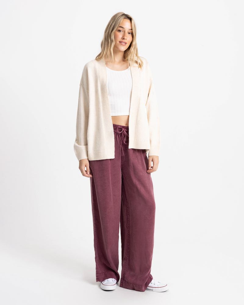 A woman wearing a Volcom Women’s Sweater Weather Cardigan Jacquard over a white cropped tank top and loose, dark red drawstring pants.