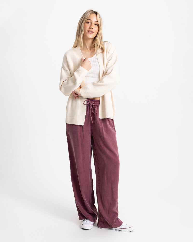 A woman wears an open-front Volcom Women’s Sweater Weather Cardigan Jacquard over a white tank top and loose-fitting maroon pants with white sneakers.