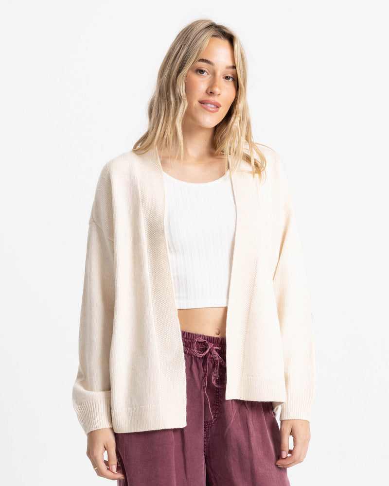 A woman wears a Volcom Women’s Sweater Weather Cardigan Jacquard in cream over a white cropped tank top and maroon drawstring pants.