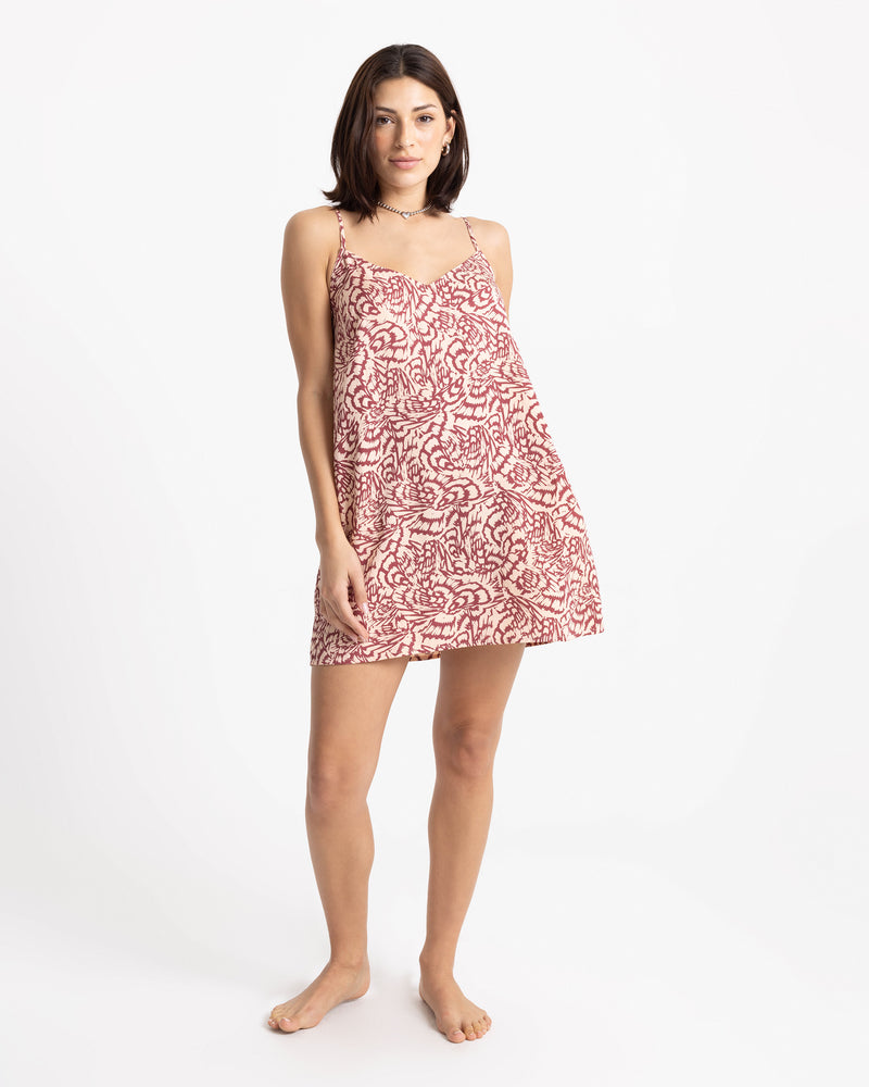 A woman wears a Volcom Women’s Boss Beach Mini Dress in a Marsala Blush colorway, featuring a sleeveless, v-neck silhouette with a red and white abstract leaf pattern.