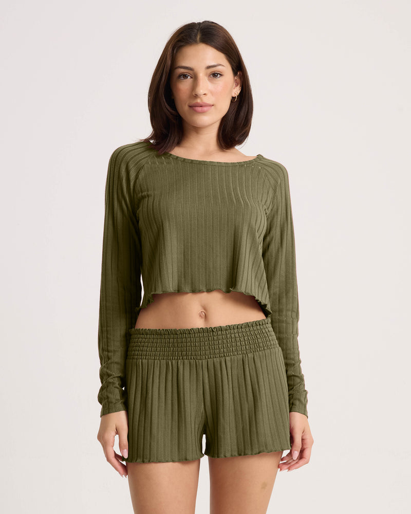 A woman with shoulder-length brown hair wears the Volcom Women’s Sun Lounge Pull Over Long Sleeve in olive green, paired with matching high-waisted shorts against a plain light background.