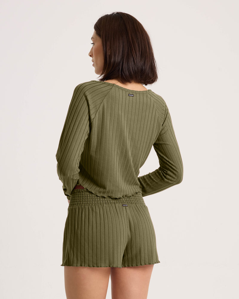 A woman with shoulder-length dark hair wears a relaxed fit, ribbed olive green Women’s Sun Lounge Pull Over Long Sleeve by Volcom and matching shorts, shown from the back against a plain light background.