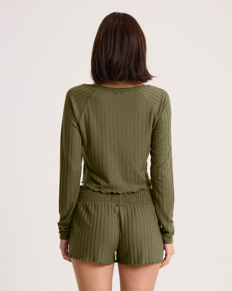 A woman with short brown hair is seen from the back wearing a relaxed fit olive green Women’s Sun Lounge Pull Over Long Sleeve by Volcom and matching shorts, posed against a plain white background.