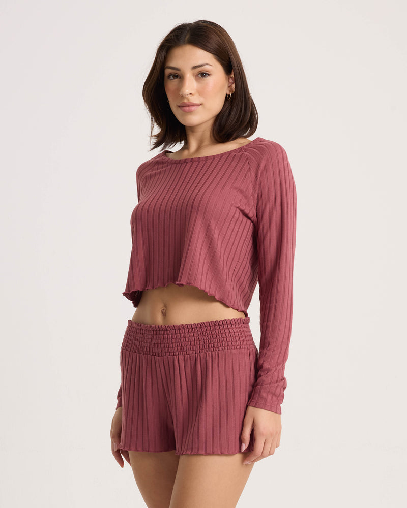 A woman wearing the Volcom Women’s Sun Lounge Pull Over Long Sleeve and high-waisted shorts stands facing forward with a neutral expression against a plain white background.
