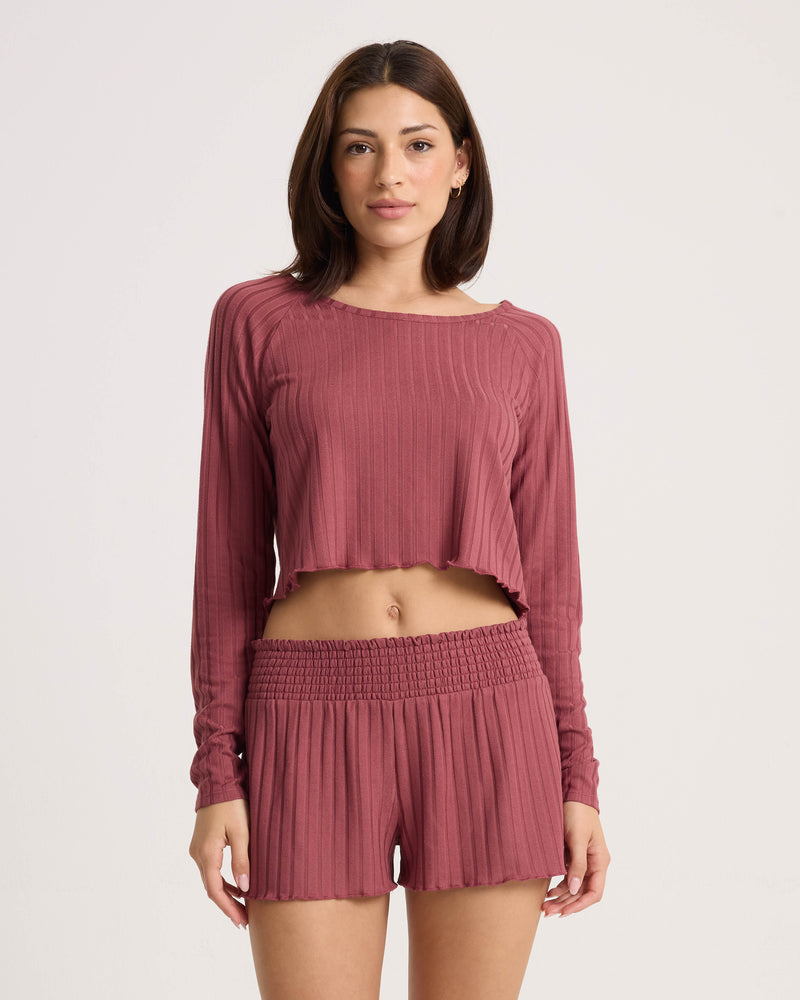 A woman faces forward in a relaxed-fit Volcom Women’s Sun Lounge Pull Over Long Sleeve and high-waisted ruffle-hem shorts, her cozy mauve set popping against a plain light background.