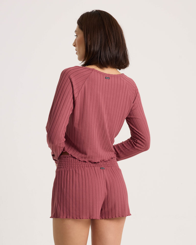 A woman with shoulder-length dark hair wears the Volcom Women’s Sun Lounge Pull Over Long Sleeve in muted pink, standing with her back to the camera against a plain background.