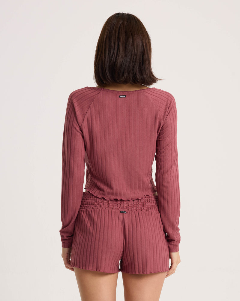 A person with shoulder-length brown hair is seen from the back, wearing a relaxed fit Women’s Sun Lounge Pull Over Long Sleeve by Volcom and matching mauve ribbed shorts, against a plain white background.