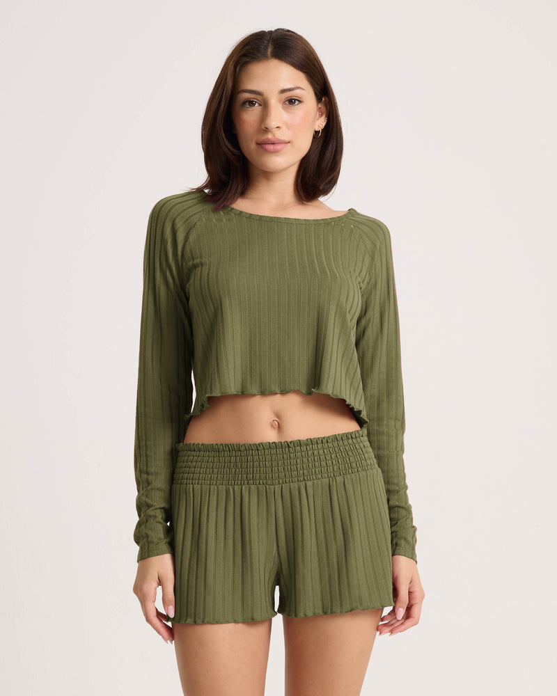 A woman stands facing forward, wearing a ribbed long-sleeve crop top and Volcom Women’s Sun Lounge Short with an elastic waistband, featuring a raw hem on the top against a plain, light-colored background.