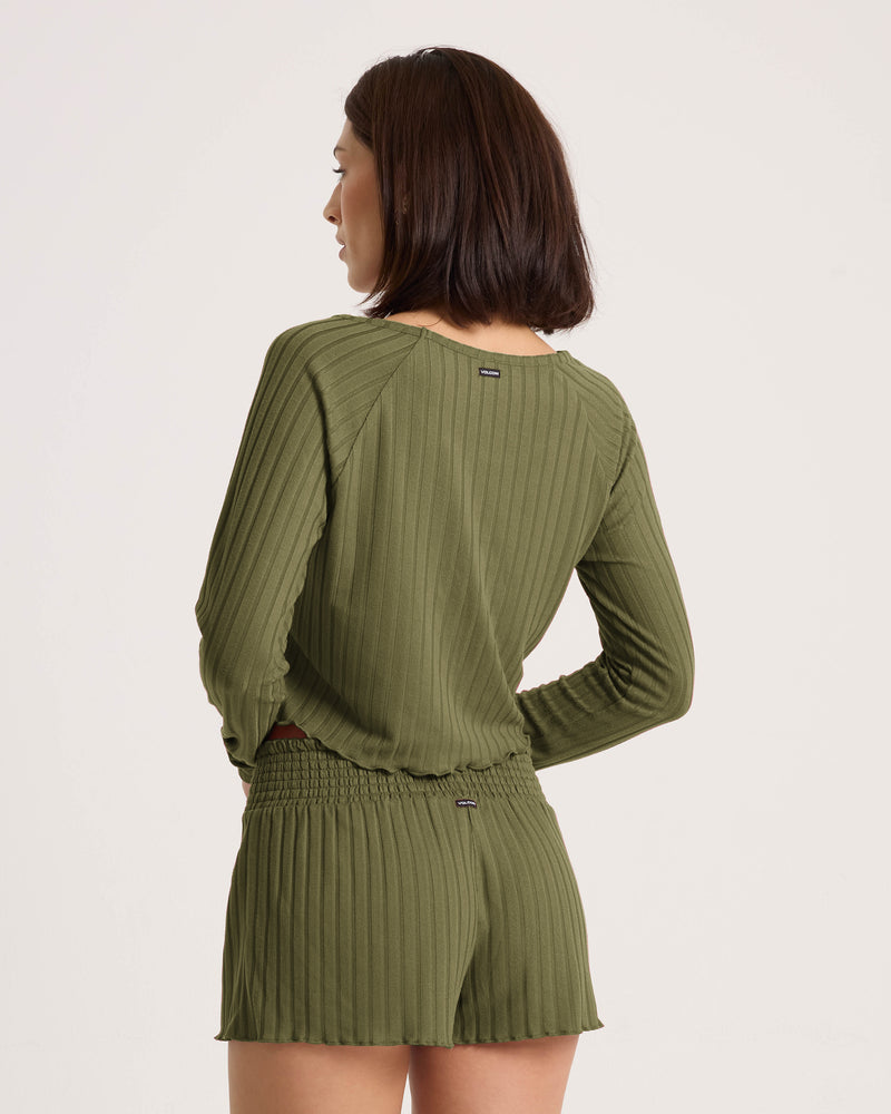 A woman with short dark hair models the Volcom Women’s Sun Lounge Short in Martini Olive with an elastic waistband, paired with a matching ribbed long-sleeve top, hands on hips against a plain background.