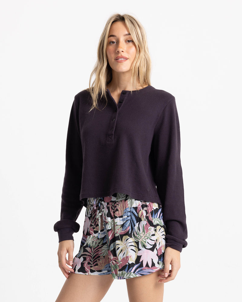 A woman wearing a dark long-sleeved henley shirt and Volcom Women’s Stone Shine Short in Garden Multi print with a tie-front drawstring waist.
