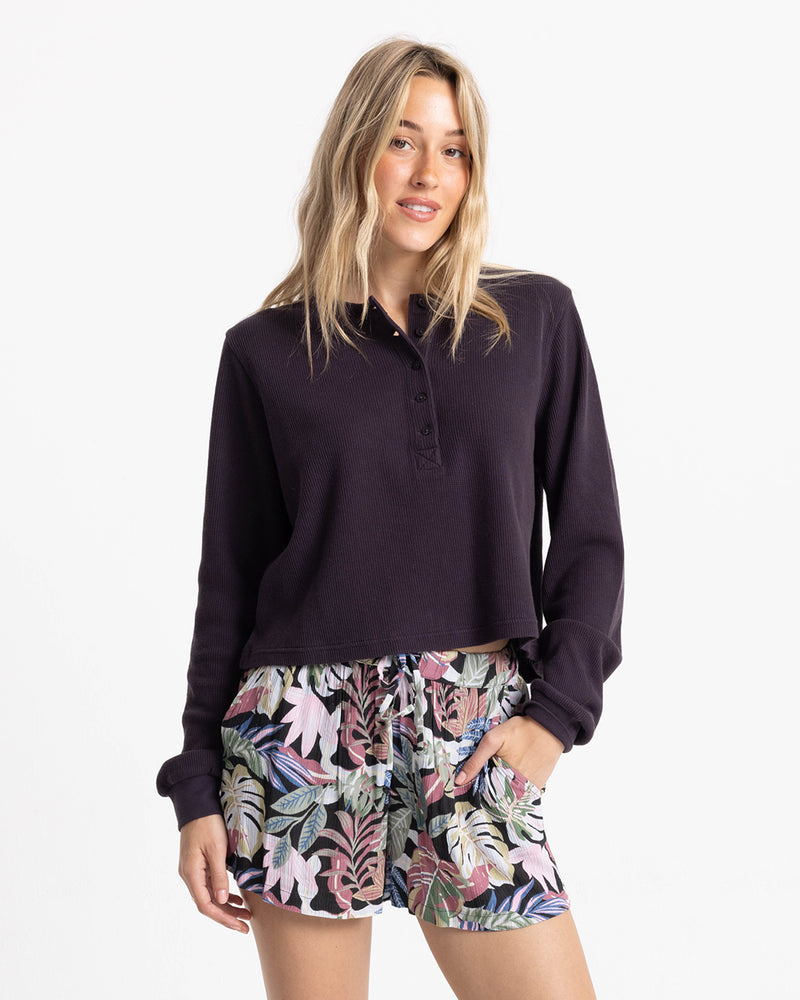 A woman wears a dark long-sleeved henley shirt and Volcom Women’s Stone Shine Short, which feature a colorful Garden Multi tropical leaf pattern and a tie-front waist.