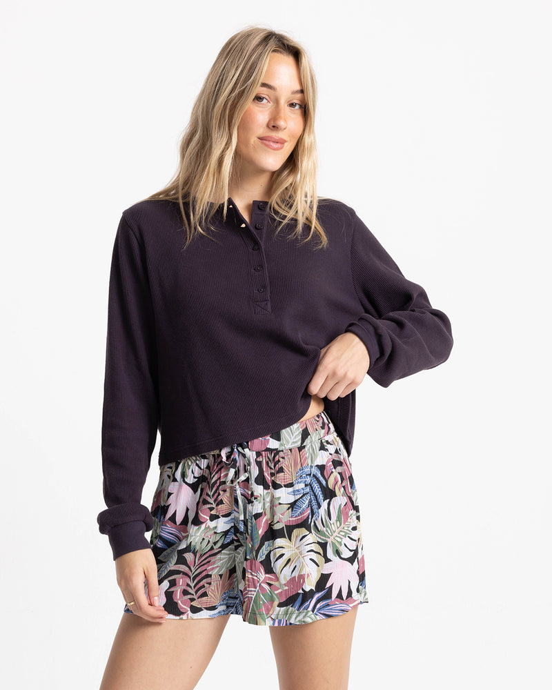 A woman wears a dark long-sleeved waffle-knit henley shirt and Volcom Women’s Stone Shine Short, which feature a tropical Garden Multi foliage pattern and a tie-front waist.
