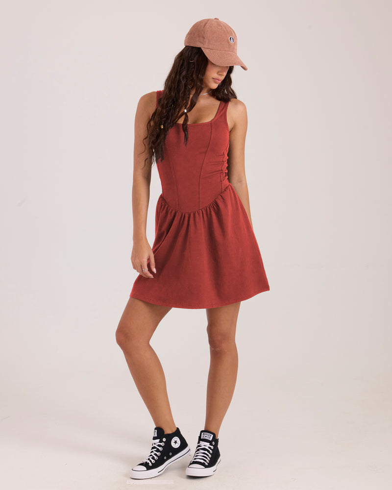 A person wearing a rust-colored Volcom Women’s Carve Dress with a dropped waist, a baseball cap, and black high-top sneakers.