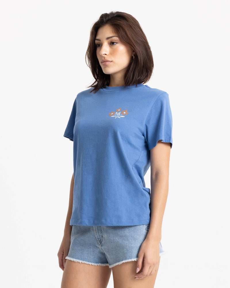 A woman wearing a Volcom Women’s Fleur De Stone Lock It Up Tee, a boxy-fit, blue short-sleeved crew neck boyfriend graphic tee with a small floral design on the left chest, paired with light-wash denim shorts.