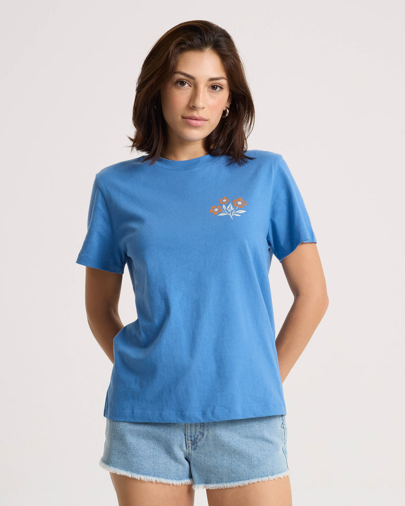 A person wearing a Volcom Women’s Fleur De Stone Lock It Up Tee, a 100% cotton blue short-sleeved t-shirt with a boxy fit and a small floral graphic on the chest.
