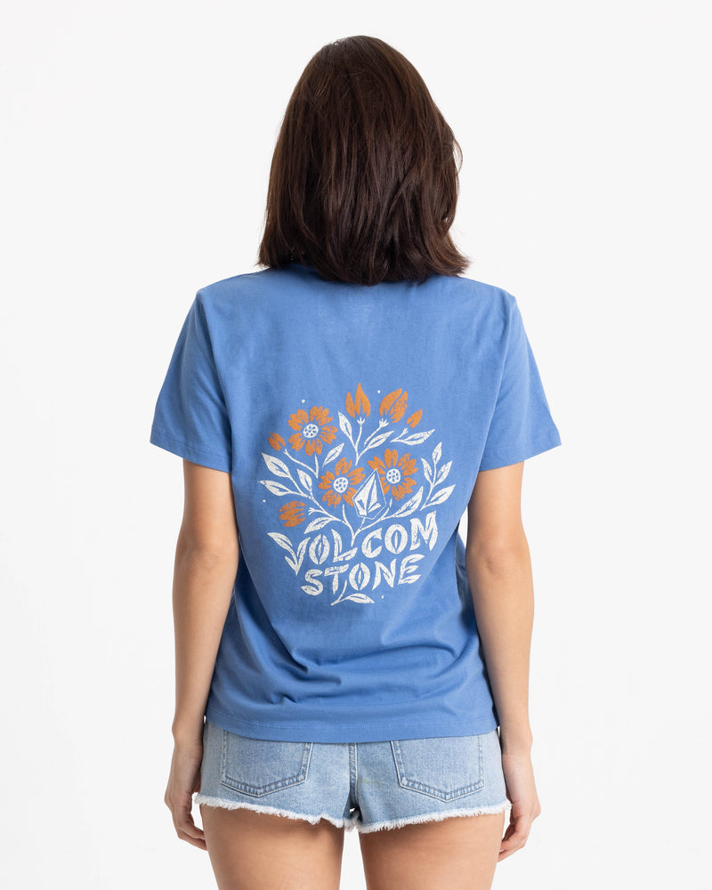 A person wearing a boxy-fit blue Women’s Fleur De Stone Lock It Up Tee with a white and orange floral graphic on the back that reads VOLCOM STONE.