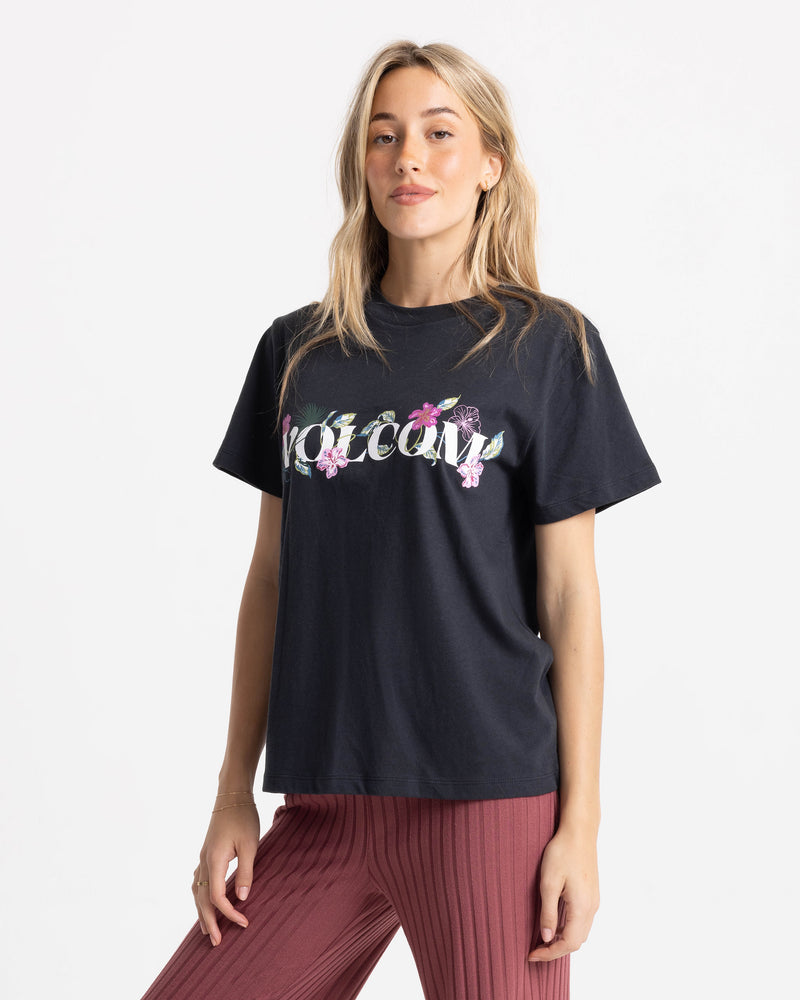 A woman in a Women’s Volcome Vines Lock ITee Up Tee with floral lettering and maroon ribbed pants stands confidently against a plain white background.