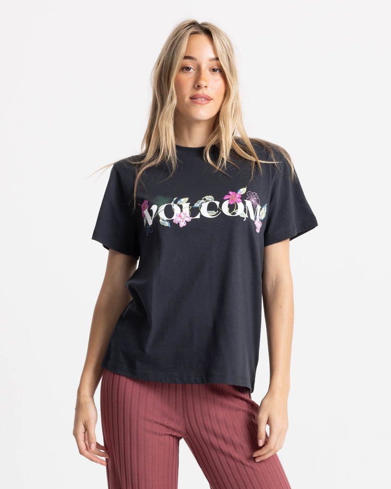 A woman with long blonde hair wears the Women’s Volcome Vines Lock ITee Up Tee, a 100% cotton black graphic tee by Volcom with WELCOME and floral designs, paired with red pants against a white background.