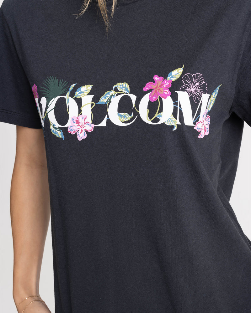 A woman wears the Women’s Volcome Vines Lock ITee Up Tee by Volcom, a black 100% cotton tee featuring VOLCOM in white with vibrant tropical flowers and leaves around the text.