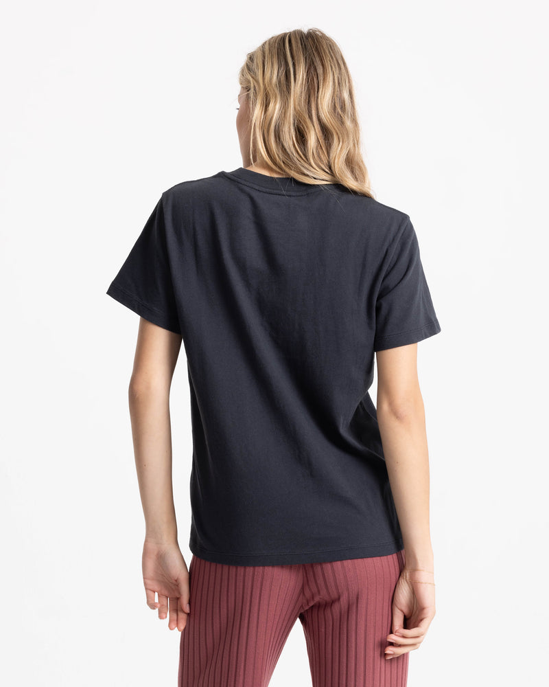 Woman with long blonde hair wears the Women’s Volcome Vines Lock ITee Up Tee by Volcom in black, paired with ribbed maroon pants, standing with her back to the camera against a plain white background.