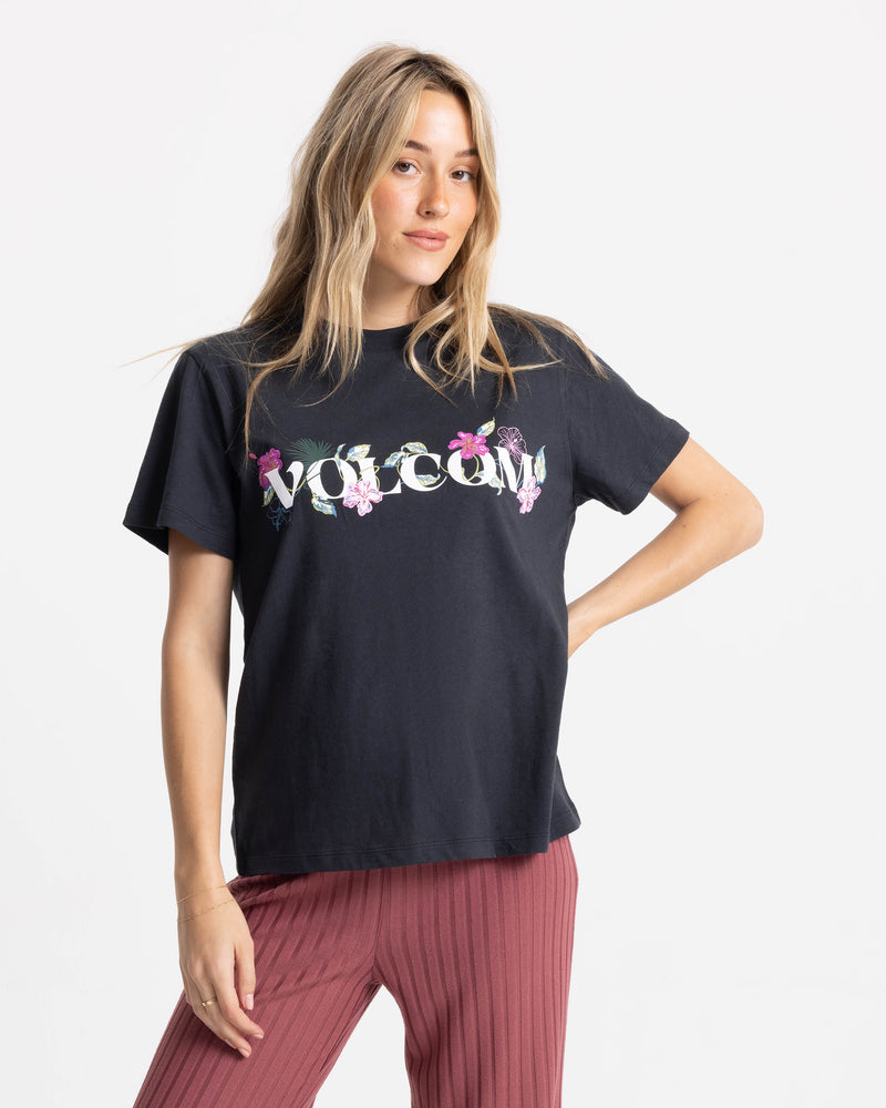 A woman with long blonde hair wears the Women’s Volcome Vines Lock ITee Up Tee by Volcom and mauve pants, posing with one hand on her hip against a plain white background.