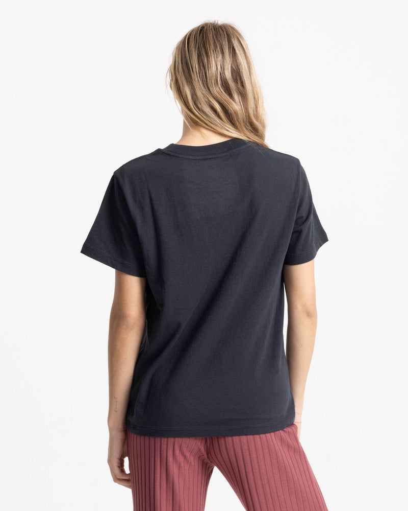 A person with long, light brown hair wears the Women’s Volcome Vines Lock ITee Up Tee and ribbed pink pants, standing with their back to the camera against a white background.