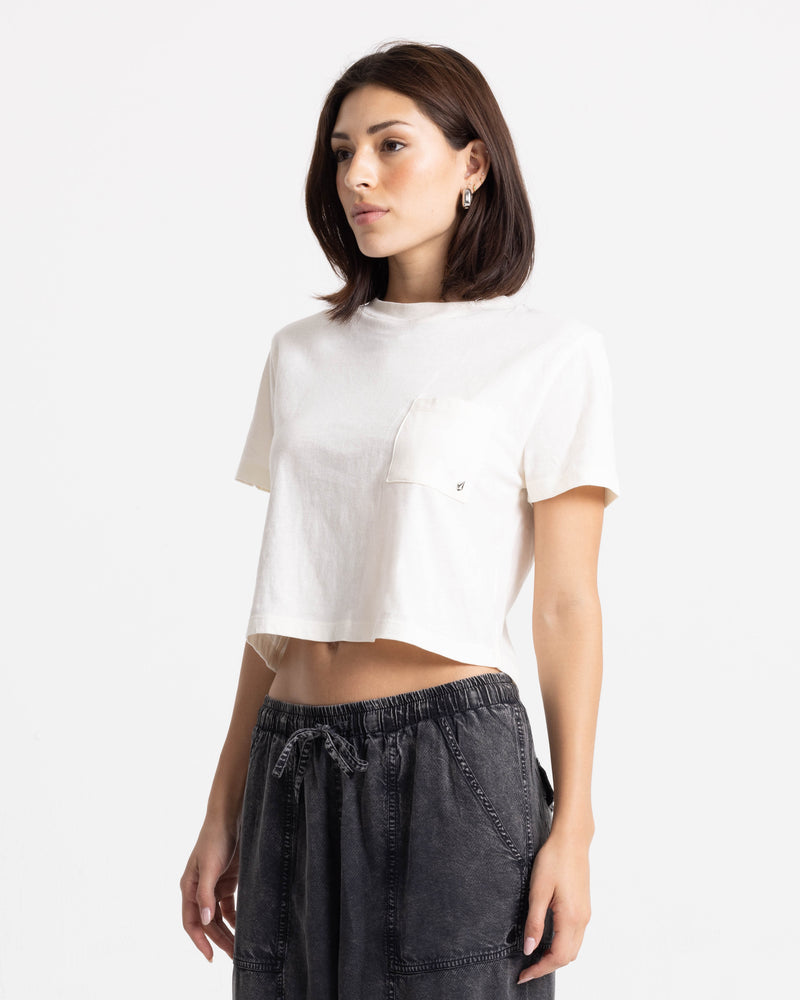 A woman with straight brown hair wears the Volcom Women’s BuTeeTeeerfly Ring Pocket Dial Tee, a 100% cotton white boxy fit shirt with a pocket, paired with dark gray drawstring pants against a plain white background.