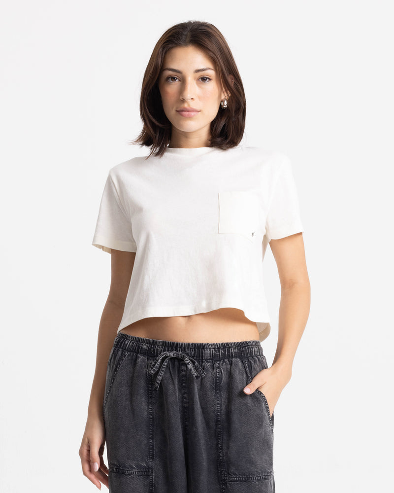 A woman wears the Volcom Women’s BuTeeTeeerfly Ring Pocket Dial Tee and black drawstring pants, standing against a white background with a neutral expression.