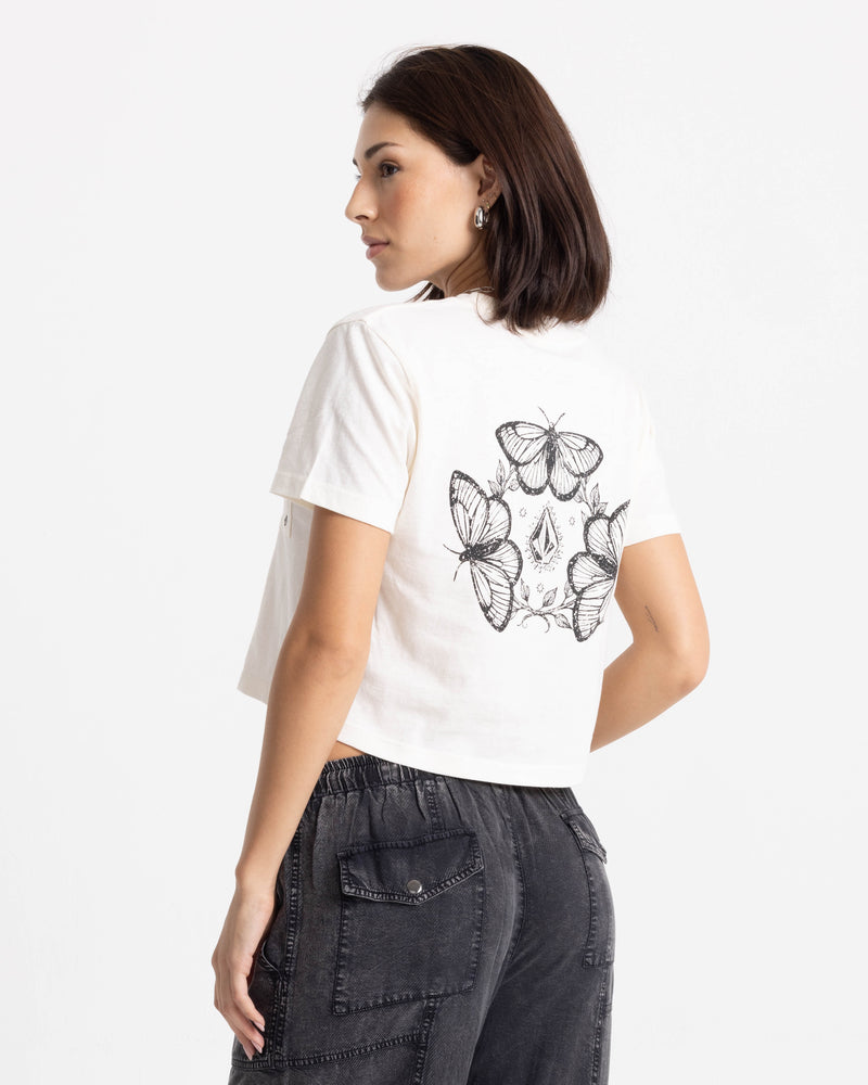A woman with shoulder-length dark hair models the Volcom Women’s BuTeeTeeerfly Ring Pocket Dial Tee—100% cotton, boxy fit with black butterfly graphics on the back—paired with dark jeans against a white background.