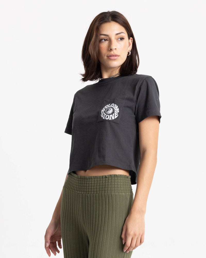 A woman with shoulder-length brown hair wears the Volcom Women’s Ying Yang Pocket Dial Tee and olive green ribbed pants, standing against a plain white background.