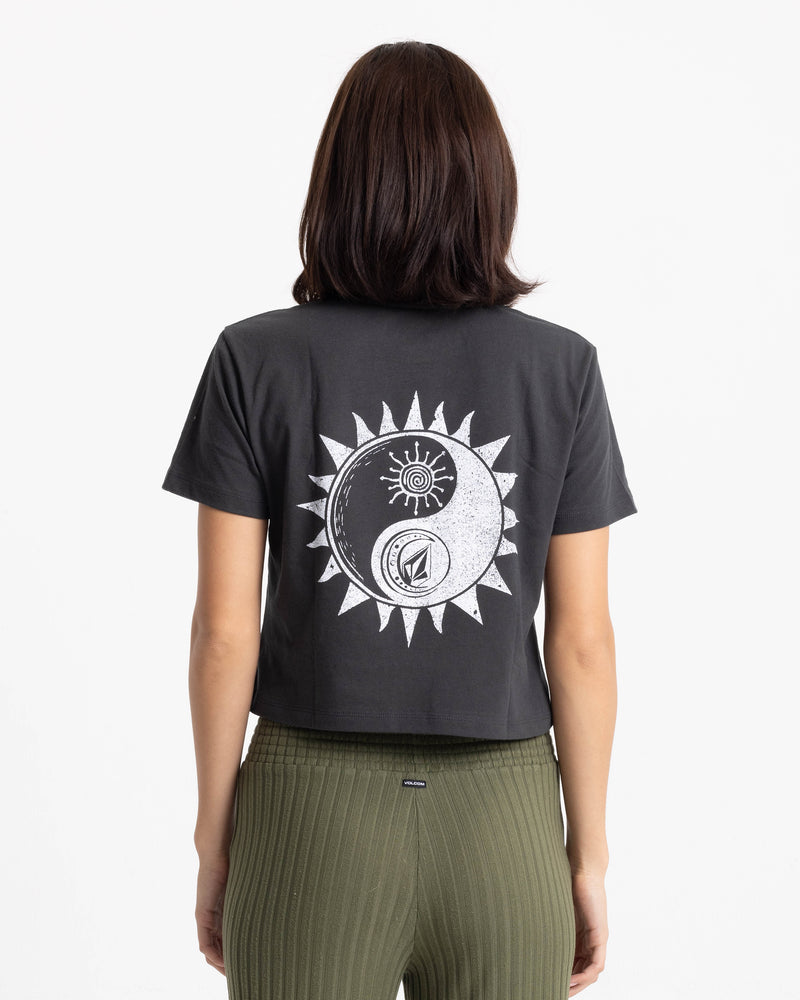 A person with shoulder-length dark hair is seen from behind wearing the Volcom Women’s Ying Yang Pocket Dial Tee, a boxy fit black T-shirt featuring white sun, moon, and yin-yang graphics on the back, paired with olive ribbed pants.
