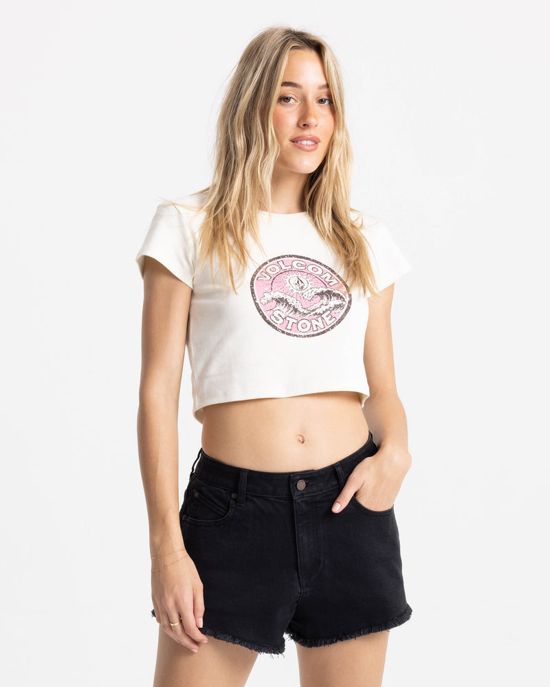 A woman with long blonde hair wears the Volcom Women’s Ride Teehe Wave 1991 Mid Tee—white, cropped, boxy fit with a pink graphic—paired with black denim shorts. She stands against a plain white background, one hand in her pocket.