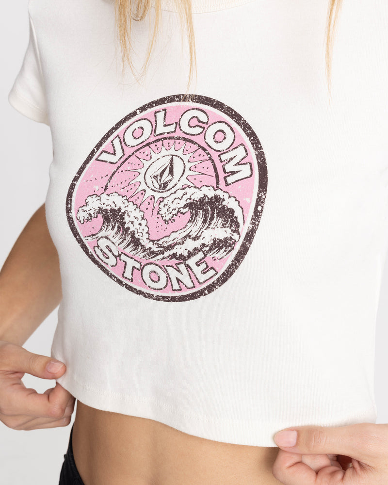 A person wears the Volcom Women’s Ride Teehe Wave 1991 Mid Tee, a white cropped boxy cotton graphic tee with pink and black wave art and the iconic stone symbol.